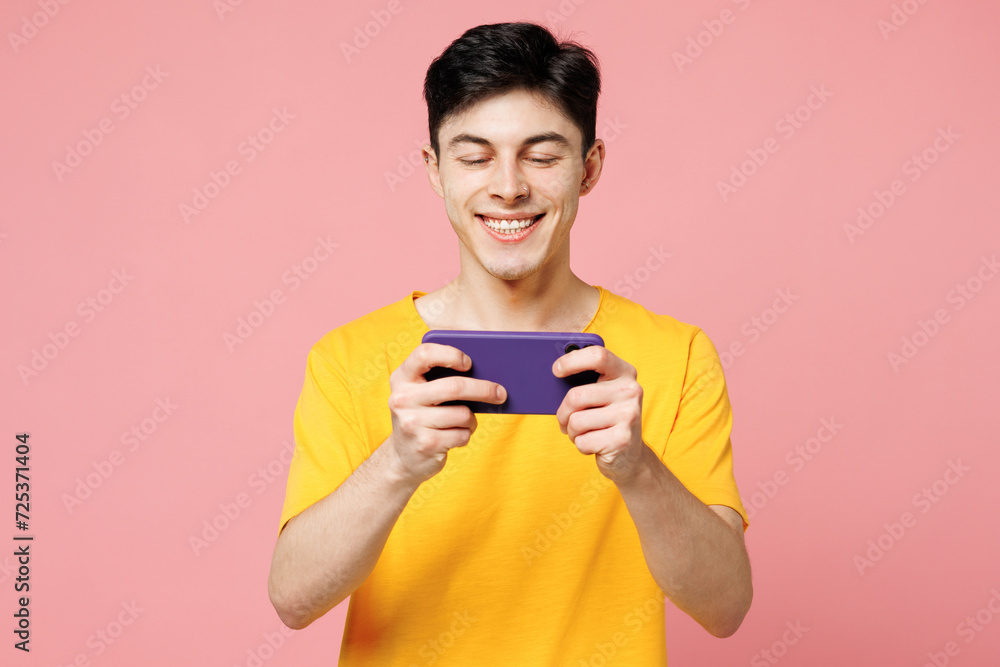 Young man he wears yellow t-shirt casual clothes use play racing app on mobile cell phone hold gadget smartphone for pc video games isolated on plain pastel light pink background. Lifestyle concept. Young man he wears yellow t-shirt casual clothes use play racing app on mobile cell phone hold gadget smartphone for pc video games isolated on plain pastel light pink background. Lifestyle concept.
