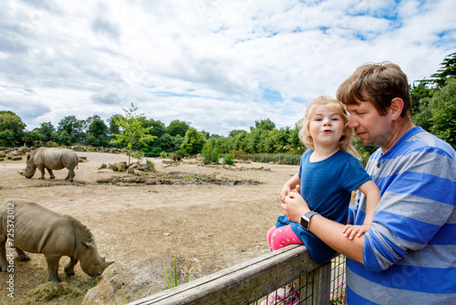 Cute adorable toddler girl and father watching wild rhinos in zoo. Happy baby child, daughter and dad, family having fun together with animals safari park on warm summer day. Ireland