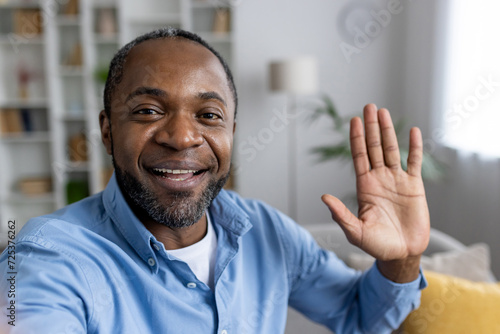 Front person view of african american man in shirt waving hello while starting video call in apartment interior. Friendly adult male greeting and welcoming interlocutor at beginning of conversation.