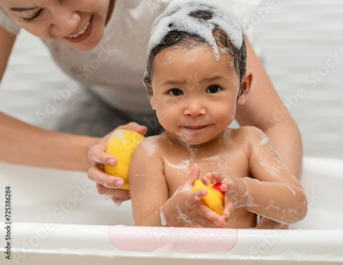 mother bathing her cheerful infant baby with a soft sponge with foam bubbles in bathtub
