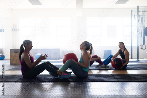 Side view of multiracial young females passing fitness balls while sitting on floor in health club