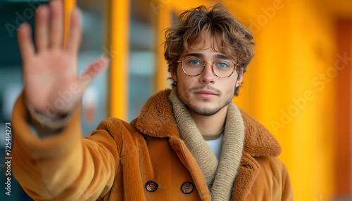 Young man in glasses and a brown coat gesturing stop with his hand, blurred background.