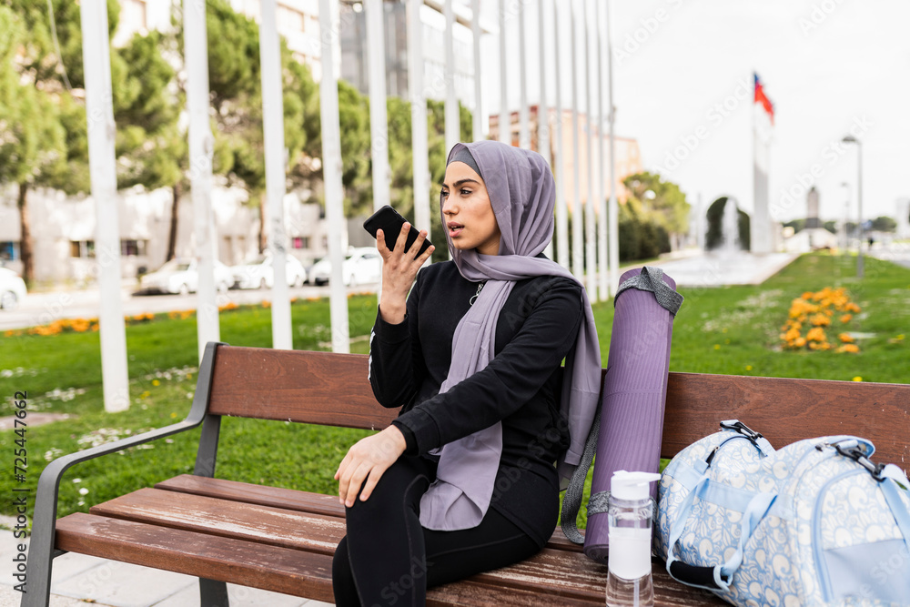 veiled woman recording audio with her cell phone while waiting for her gym partner sitting on a bench veiled woman recording audio with her cell phone while waiting for her gym partner sitting on a bench
