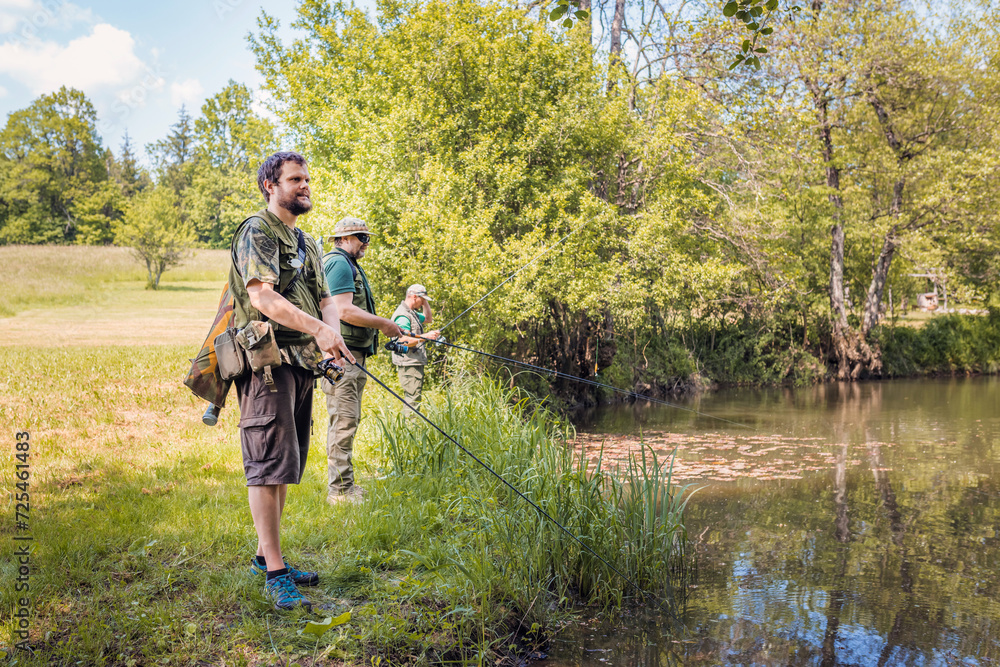 Three sports fishermen standing on the river bank, under a beautiful tree, swinging their fishing rods trying to catch fish on a sunny summer day. Three sports fishermen standing on the river bank, under a beautiful tree, swinging their fishing rods trying to catch fish on a sunny summer day.