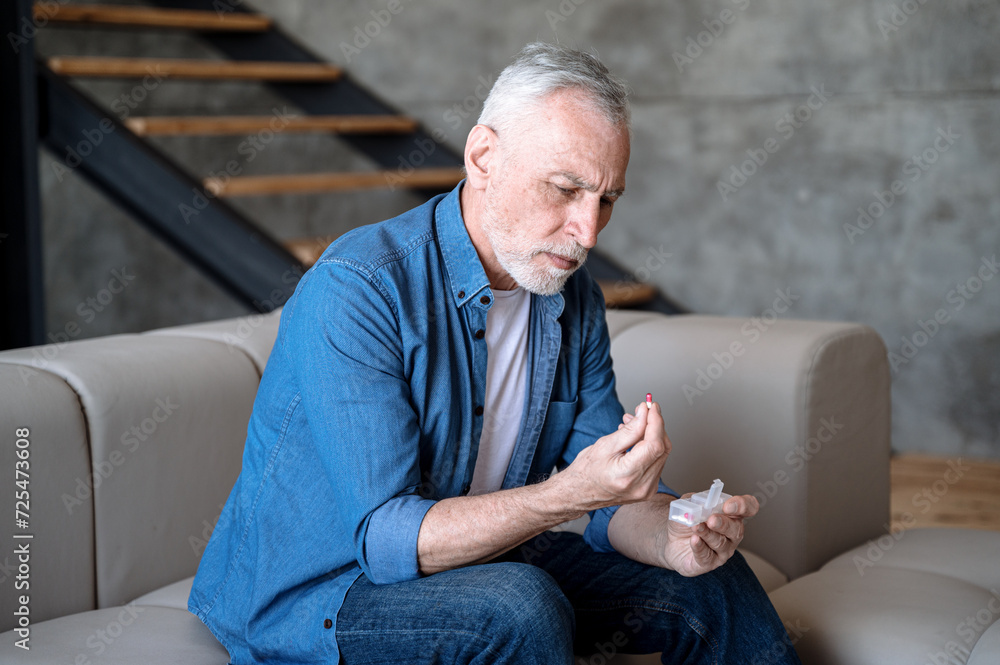 Senior man sitting on sofa and holding pill from daily organizer. Senior man sitting on sofa and holding pill from daily organizer.