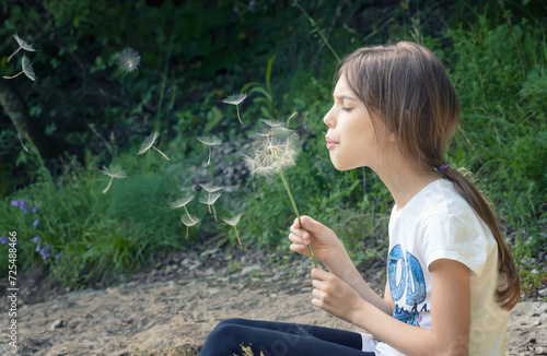 Closeup portrait of a beautiful little girl blowing dandelion on the background of a green field
