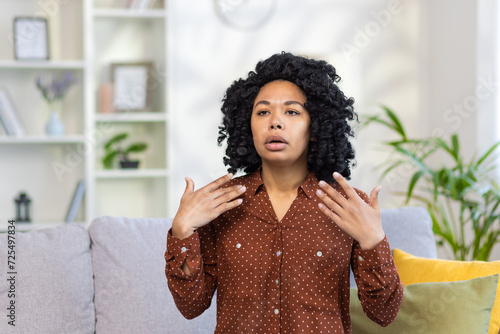 African-American young woman is sitting on the sofa at home where it is very hot, she is not feeling well, she is waving her hands in her face, she needs fresh air