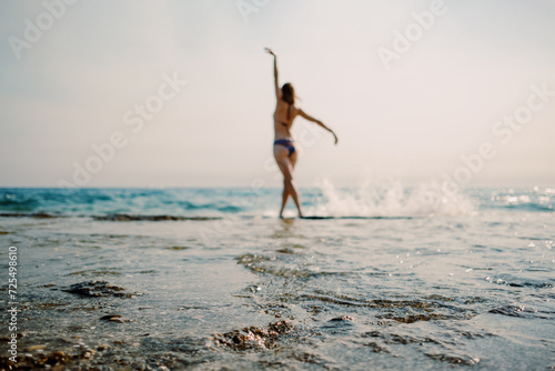 A woman with arms raised in a moment of freedom at the sunlit seashore, with gentle waves lapping at her feet