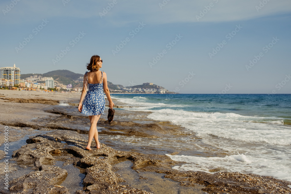 A carefree woman takes a leisurely stroll along the seaside in a flowing summer dress, with the Mediterranean landscape in the background