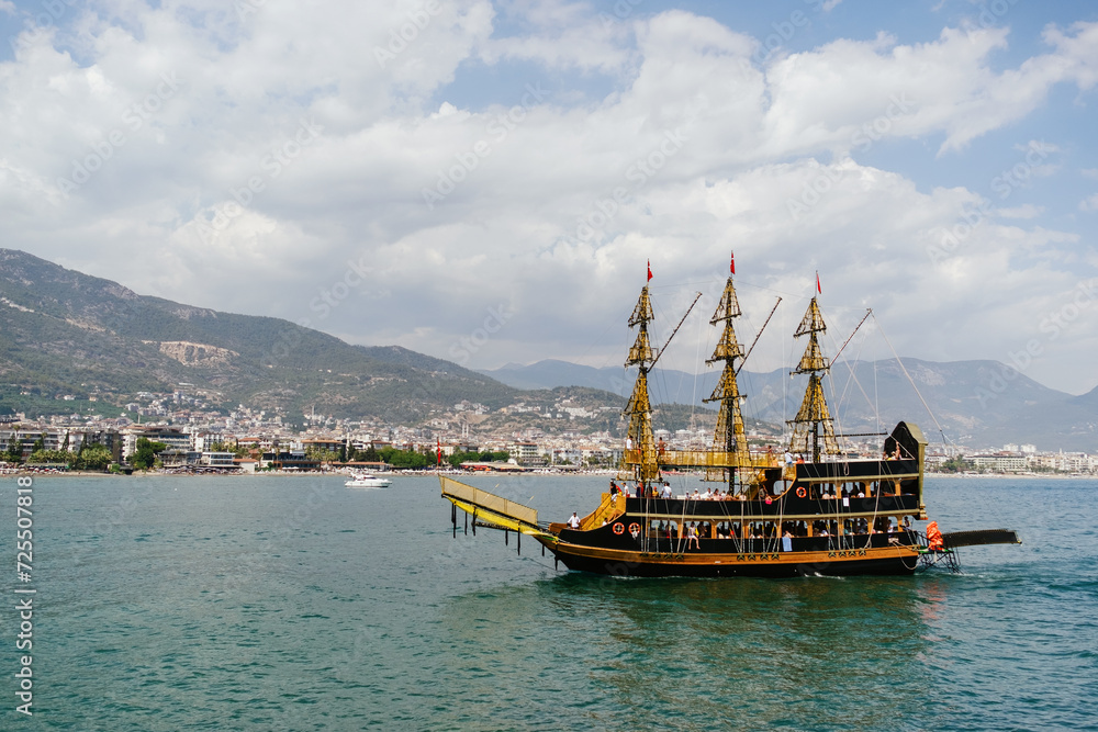 A pirate-themed tour boat sails across a calm Mediterranean bay with a scenic coastal town and mountains in the background A pirate-themed tour boat sails across a calm Mediterranean bay with a scenic coastal town and mountains in the background
