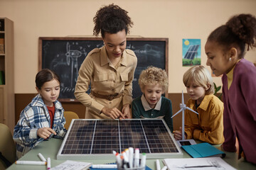 Diverse group of children learning about renewable energy in school with young teacher sowing solar panel to class