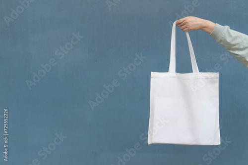 Blank white tote bag canvas fabric with handle mock up design. Close up of woman hand holding eco or reusable shopping bag on blue metal wall. No plastic bag and ecology concept.