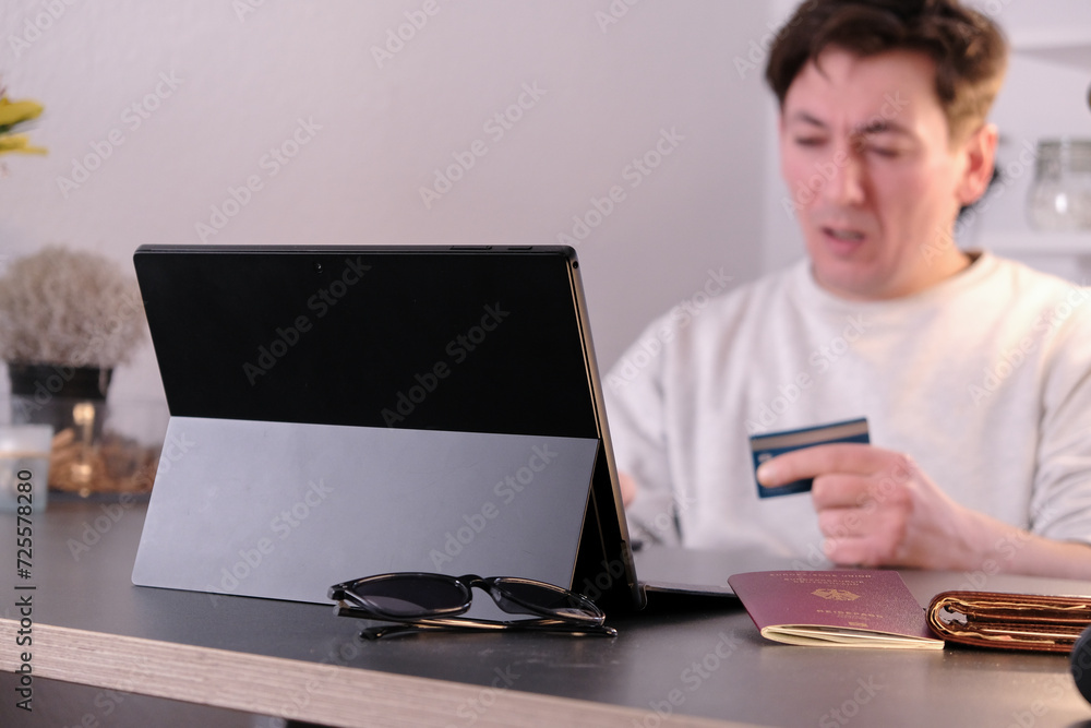 Shocked young man with a laptop and credit card in hand, passport and wallet on the table Shocked young man with a laptop and credit card in hand, passport and wallet on the table