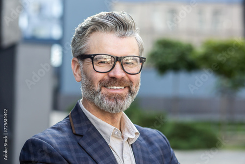 Portrait of a cheerful senior male professional with gray hair, wearing glasses and a stylish business suit outside.