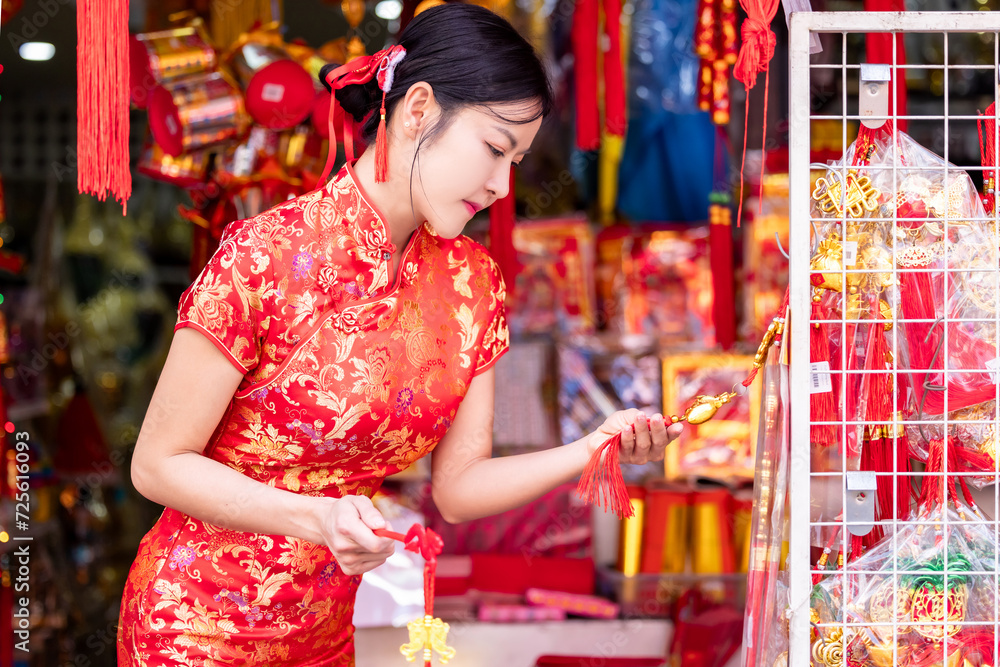 Asian woman in red cheongsam dress choosing and buying home decoration and ornament for celebrating Chinese Lunar New Year festival at market.Celebrate Chinese lunar new year.Celebrate season holiday. - obrazy, fototapety, plakaty 