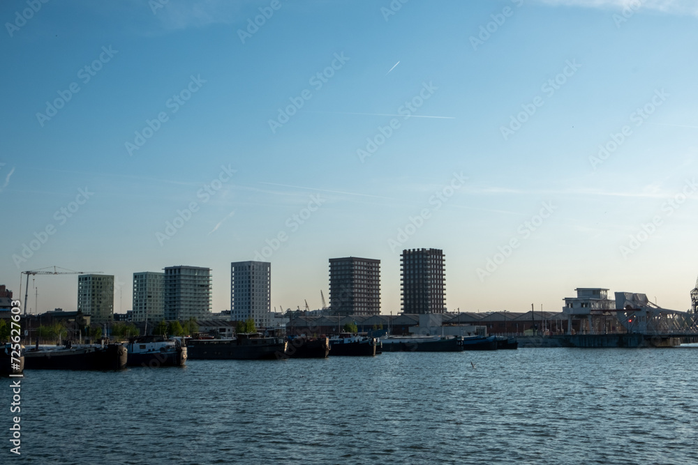 Antwerp, 20th of April, 2022, The photograph captures a late afternoon at the evolving waterfront of Antwerp. The image is dominated by the port's waters in the foreground, with a variety of moored