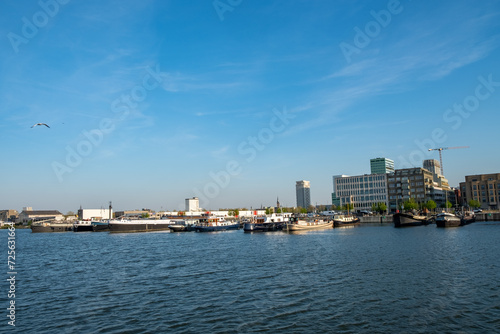 Antwerp, 20th of April, 2022, This image presents a lively scene at the Port of Antwerp. The waters in the foreground are dotted with boats and ships, implying a busy maritime hub. Across the water