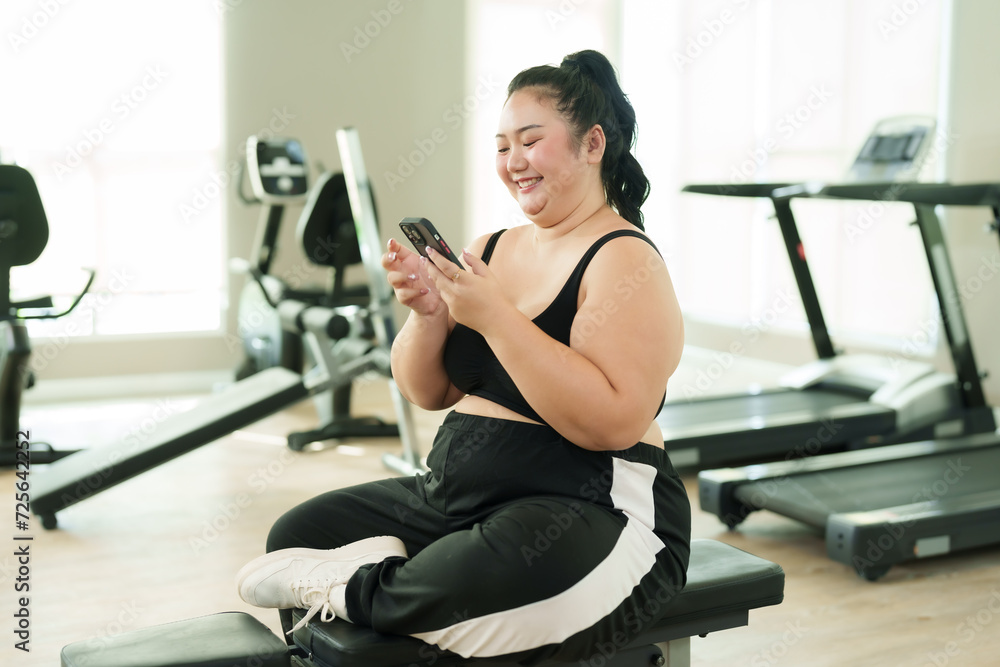 Overweight Asian woman exercise in gym, Joyful in activewear using smartphone, resting on bench with treadmills in behind. Smiling plus-sized female casually seated on bench, itness machines behind Overweight Asian woman exercise in gym, Joyful in activewear using smartphone, resting on bench with treadmills in behind. Smiling plus-sized female casually seated on bench, itness machines behind