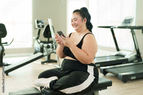 Overweight Asian woman exercise in gym, Joyful in activewear using smartphone, resting on bench with treadmills in behind. Smiling plus-sized female casually seated on bench, itness machines behind