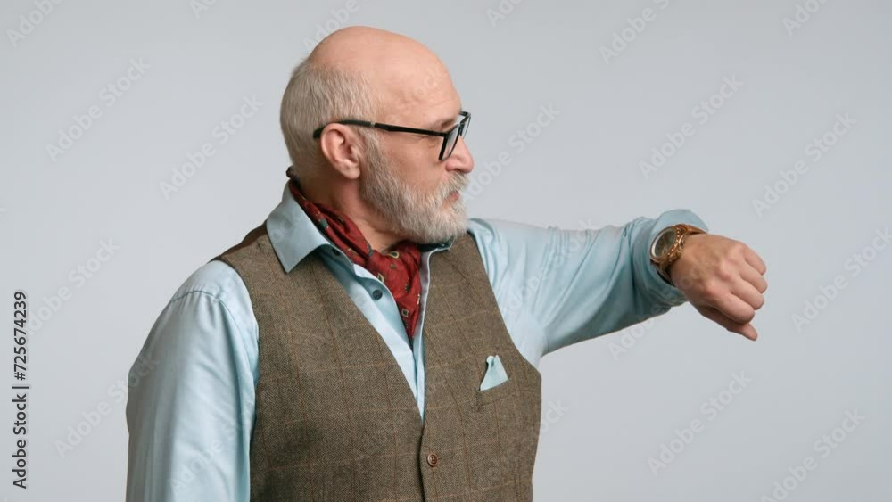 An authoritative senior in a tailored vest makes a point by indicating his wristwatch, symbolizing the value of time, posed against a minimalist light background. High quality 4k footage An authoritative senior in a tailored vest makes a point by indicating his wristwatch, symbolizing the value of time, posed against a minimalist light background. High quality 4k footage