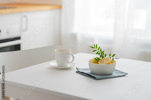 A cup of tea with lemon and a teapot on a white table against the background of a white kitchen with wooden countertop.