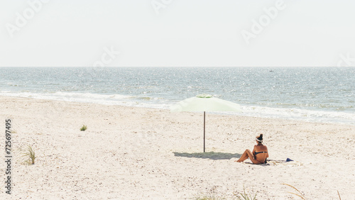 Fototapeta Naklejka Na Ścianę i Meble -  Rear view of a young woman sitting with an umbrella on the beach during a summer vacation