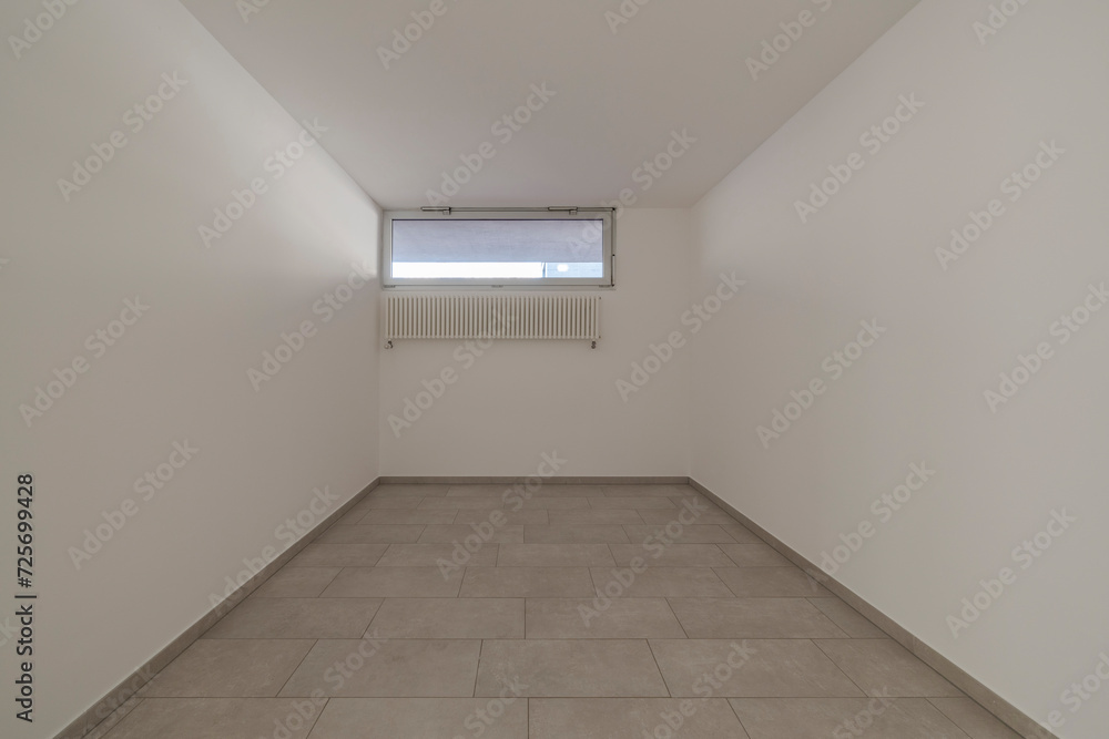 Front view of a small empty bedroom with white walls a small window and the radiator hanging on the wall. Front view of a small empty bedroom with white walls a small window and the radiator hanging on the wall.