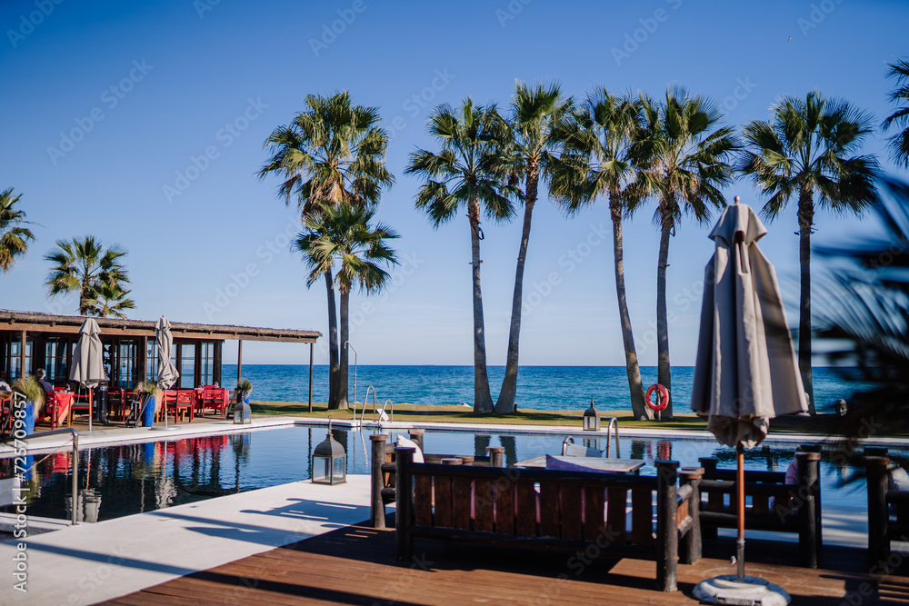 Sotogrante, Spain - January 25, 2024 - A seaside infinity pool lined with palm trees and red chairs, overlooking a clear blue ocean under a bright sky.