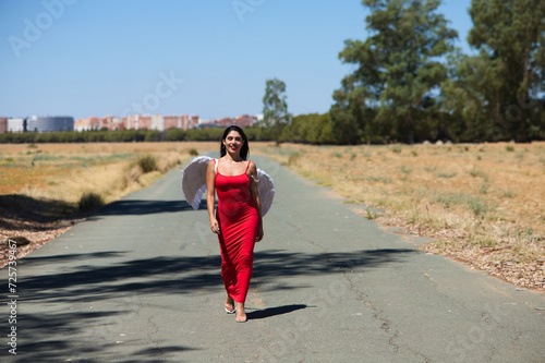 Beautiful young Latin woman in red silk dress and white wings walks along a tarmac road in the countryside outside the big city. The woman makes different poses and expressions.