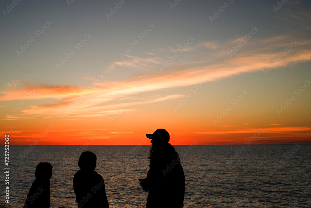 Orange sky background with clouds at sunset and black silhouettes of people against the sky and sea on a summer evening. Seascape