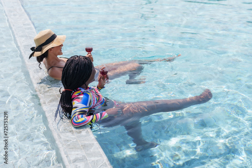 Two women are close friends different races, hispanic with African American, standing on edge swimming pool, holding glass wine, chatting happily, their faces showed that they were extremely happy.