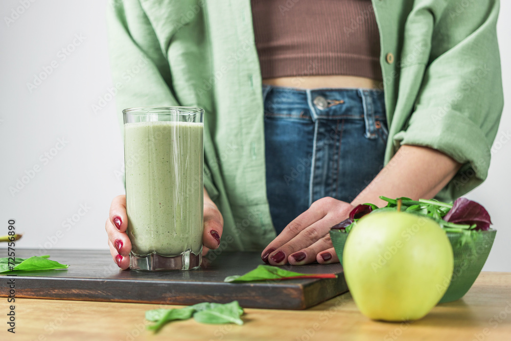 Unrecognizable woman in casual wear holding glass of fresh blended healthy green vegan smoothie. banana, spinach, apple and avocado on a table Unrecognizable woman in casual wear holding glass of fresh blended healthy green vegan smoothie. banana, spinach, apple and avocado on a table