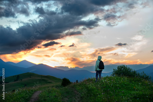 A girl tourist admires the sunset in the mountains. Silhouettes of mountains against the background of the sunset sky and a dirt road through a green meadow with wild flowers.