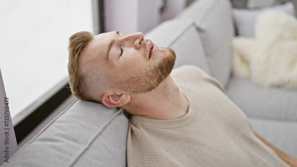 A handsome caucasian man with a beard relaxes on a modern couch in a well-lit, cozy living room A handsome caucasian man with a beard relaxes on a modern couch in a well-lit, cozy living room