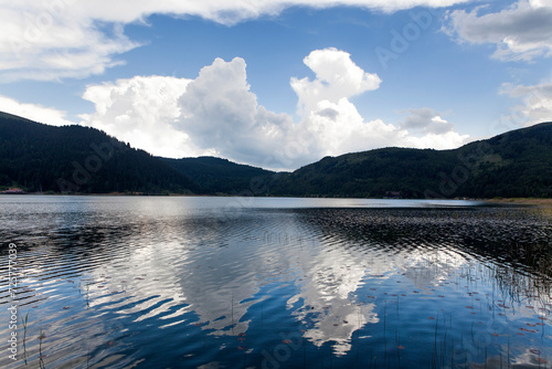 Wide scenic view of a beautiful mountain lake with dramatic white clouds and blue sky reflecting perfectly in the calm water surface.