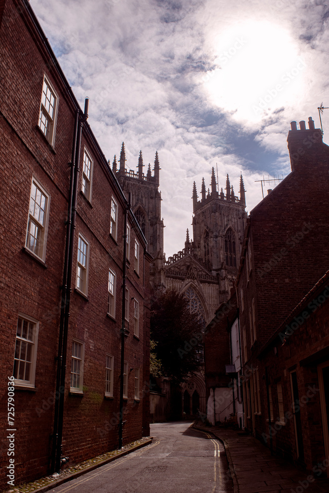 Medieval York Majesty: Sunlit Alley, York Minster Peeking Behind Houses, Red Brick Charm, Tranquil Urban Scene with Blue Sky – A Timeless Glimpse of Historic England