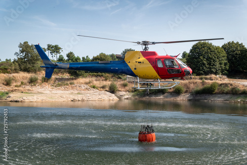 Operação aérea de combate: Helicóptero bombeiro recarrega o seu balde suspenso em lago durante intervenção contra um incêndio florestal photo