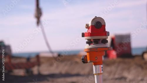 Close-up image of optical level or surveying instrument used to measure angles and distances on construction site. In background, construction worker using crane to lift materials and bulldozer . sea photo