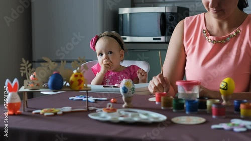 Adorable baby girl in pink dress eating cookie while her mother painting Easter egg at table in the kitchen at home photo