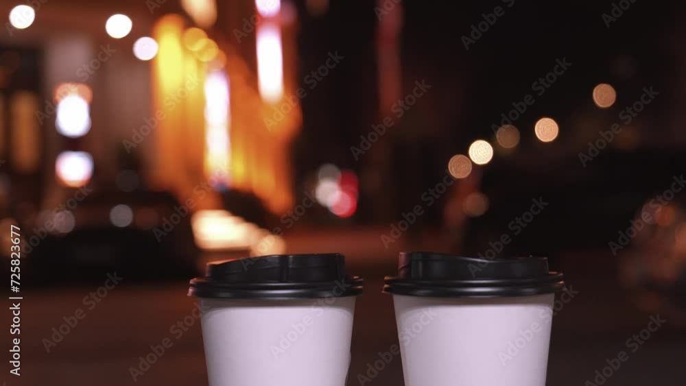 Two white paper disposable cups on blurred background of evening city lights. Female unrecognizable hand taking one cup of coffee. Hot Takeaway Coffee Outdoors. Street food, Two white paper disposable cups on blurred background of evening city lights. Female unrecognizable hand taking one cup of coffee. Hot Takeaway Coffee Outdoors. Street food,