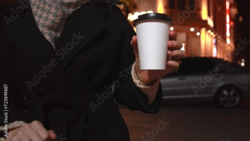 Two disposable cups made of white paper on blurred background of evening city lights. Woman taking one cup of coffee from parapet and drinking. Hot Takeaway Coffee Outdoors. Street food, photo