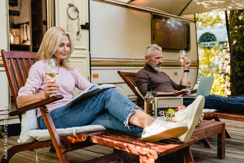 Beautiful mature blond woman reading and drinking wine in front of the motor home while her husband sitting on the deck chair using laptop for remote work, freelance, online shopping