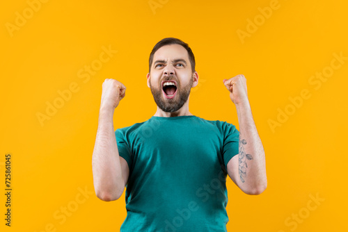 Joyful, excited, glad bearded man with wide open mouth and clenched fists celebrating money win in online lottery or at bookmaker's, isolated over yellow background photo