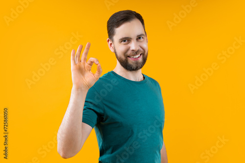Studio shot of attracting smiling young man showing ok gesture, isolated over bright colored orange yellow background
