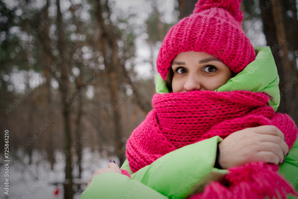 A beautiful woman in a green jacket and a pink hat and scarf is frozen and trying to keep warm standing in a winter forest A beautiful woman in a green jacket and a pink hat and scarf is frozen and trying to keep warm standing in a winter forest