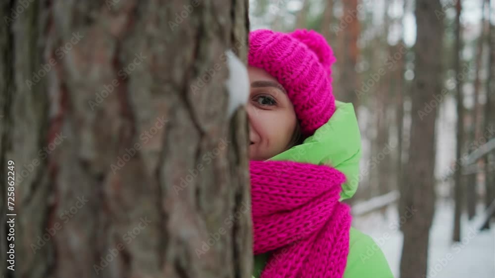 A beautiful woman in a green jacket and a pink hat and scarf hides behind a tree in a winter forest A beautiful woman in a green jacket and a pink hat and scarf hides behind a tree in a winter forest