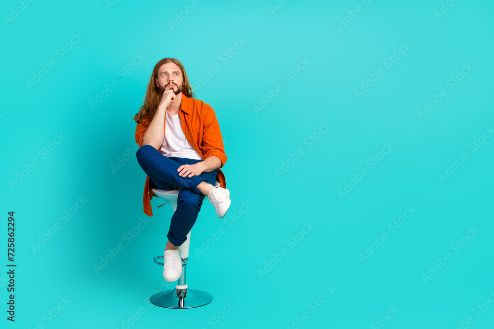 Full length photo of clever man dressed orange shirt sit on chair hand on chin look at offer empty space isolated on teal color background Full length photo of clever man dressed orange shirt sit on chair hand on chin look at offer empty space isolated on teal color background
