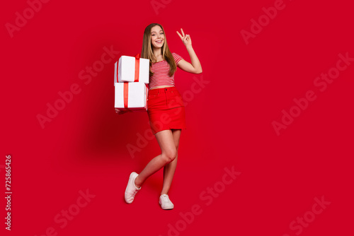Full length photo of positive cheerful lady dressed top mini skirt holding present boxes showing v-sign empty space isolated red color background