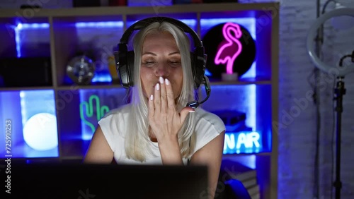 Sexy young blonde gamergirl confidently blows a kiss at the camera while gaming, turning the dark room into a fun space of love and joy. photo