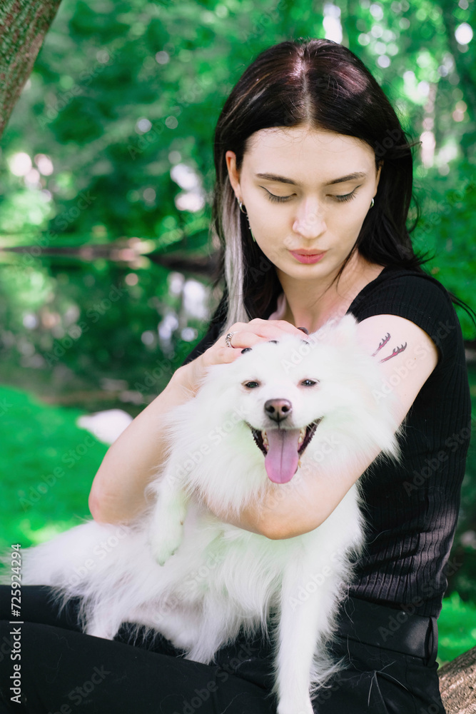 Brunette young girl in black clothes walking in the green par with a white pomeranian funny dog Brunette young girl in black clothes walking in the green par with a white pomeranian funny dog